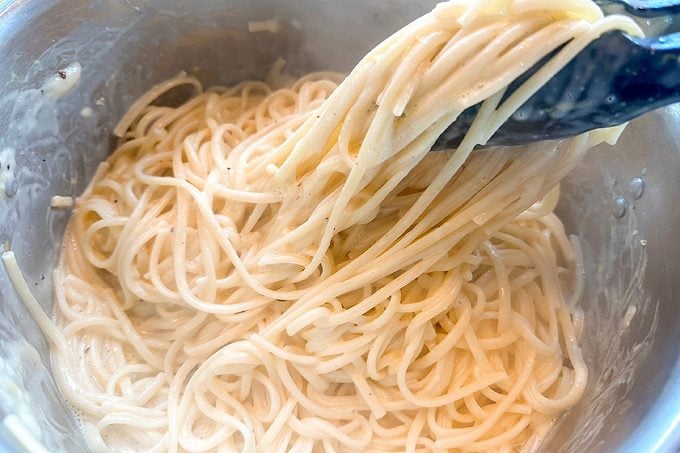 Close-up of creamy spaghetti noodles being lifted with black tongs from a metal bowl, creating an appetizing, warm, homemade cooking scene.