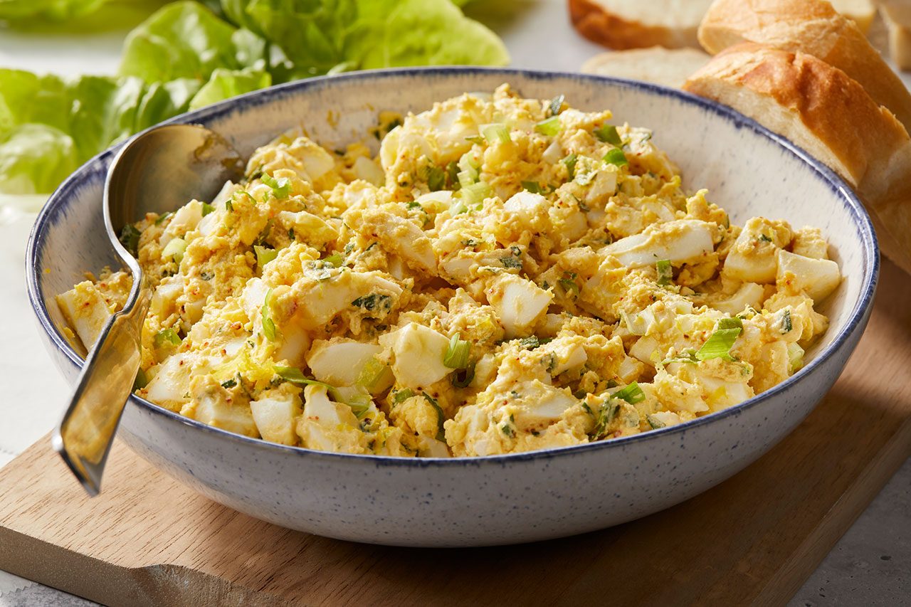 Eye level horizontal image of a large speckled blue and white bowl filled with creamy egg salad, garnished with chopped green onions. A serving spoon rests in the salad, with fresh lettuce and sliced bread visible in the soft background.