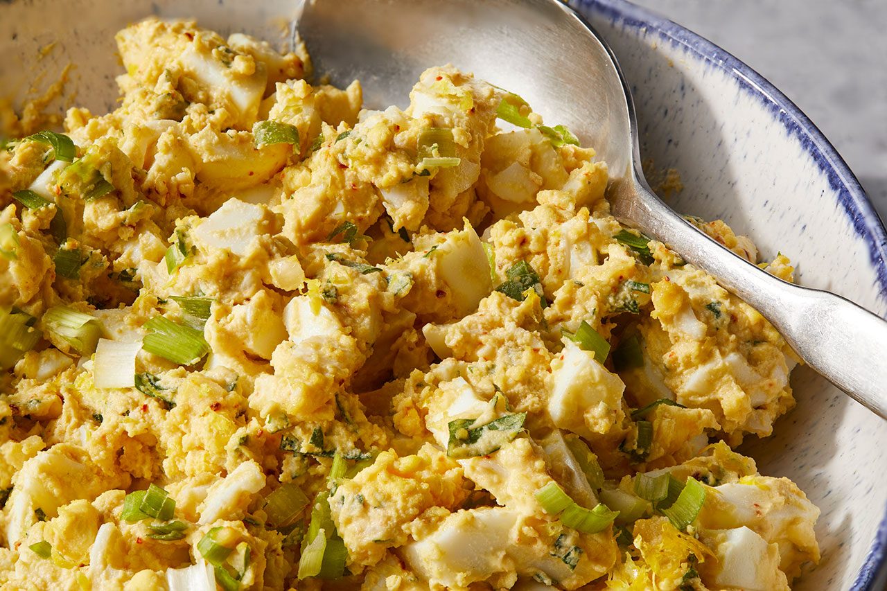 Eye Level horizontal image of a close-up of freshly made egg salad served in a decorative bowl. The creamy yellow egg salad features chopped hard-boiled eggs, vibrant green onions, and hints of red spice, with a silver spoon resting within the mixture.