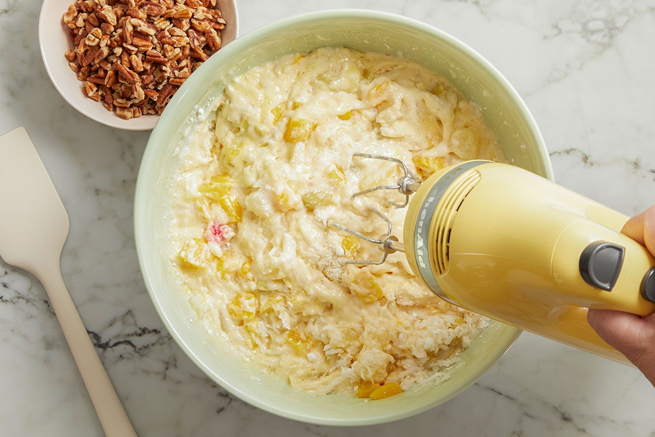 Overhead horizontal image of a hand using a yellow KitchenAid hand mixer to combine ingredients in a light green mixing bowl. The mixture contains diced yellow fruit, shredded coconut, and small red fruit pieces, with a bowl of chopped pecans and a white spatula visible on the marble surface.