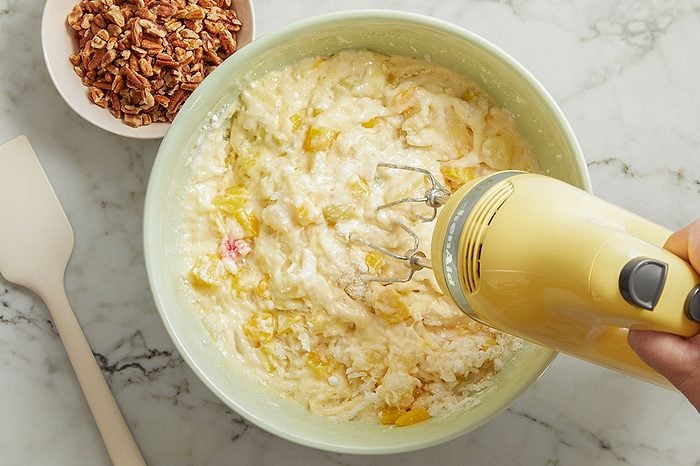Overhead horizontal image of a hand using a yellow KitchenAid hand mixer to combine ingredients in a light green mixing bowl. The mixture contains diced yellow fruit, shredded coconut, and small red fruit pieces, with a bowl of chopped pecans and a white spatula visible on the marble surface.