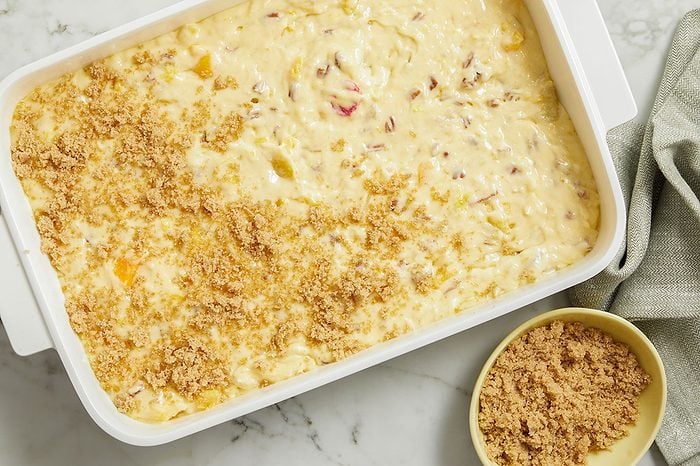 Overhead horizontal image of a white rectangular baking dish partially covered with a brown sugar topping. The dish contains a creamy, light-colored batter with visible pieces of fruit and chopped pecans. A small yellow bowl filled with brown sugar and a grey napkin are positioned next to the dish on a white marble surface.