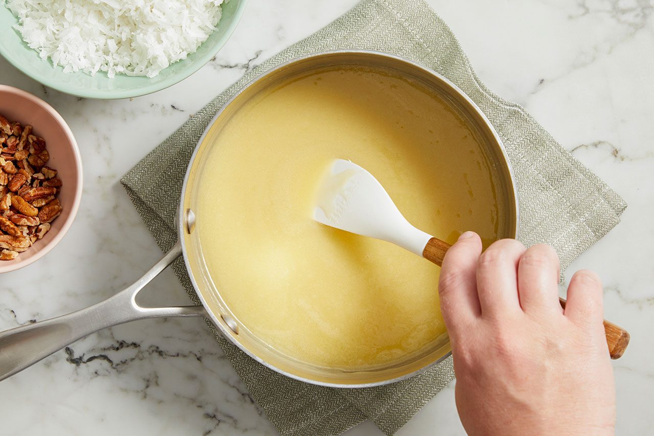 Overhead horizontal image of a hand stirring a light yellow mixture in a silver saucepan with a white silicone spatula. The saucepan rests on a light green napkin on a marble countertop, with bowls of shredded coconut and chopped pecans visible.