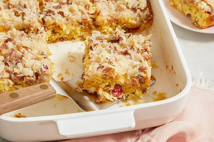 Overhead horizontal image of a piece of cake being lifted from a white rectangular baking dish with a metal spatula. The golden cake is topped with a sweet coconut and pecan frosting, with bits of red candied fruit or cherries visible within the cake. Other cake pieces remain in the baking dish.